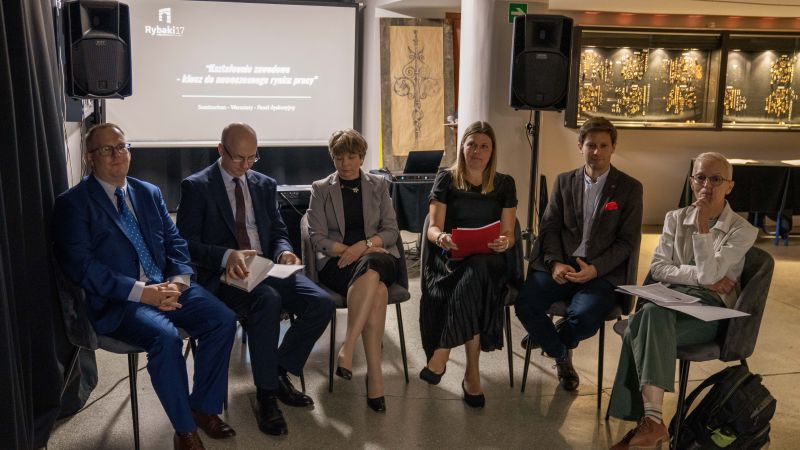 Sześć osób siedzących na krzesłach w rzędzie podczas wydarzenia "Kształcenie zawodowe - klucz do nowoczesnego rynku pracy". W tle ekran z prezentacją. Uczestnicy trzymają notatki, obok sprzęt nagłaśniający oraz gabloty zawierające biżuterię.