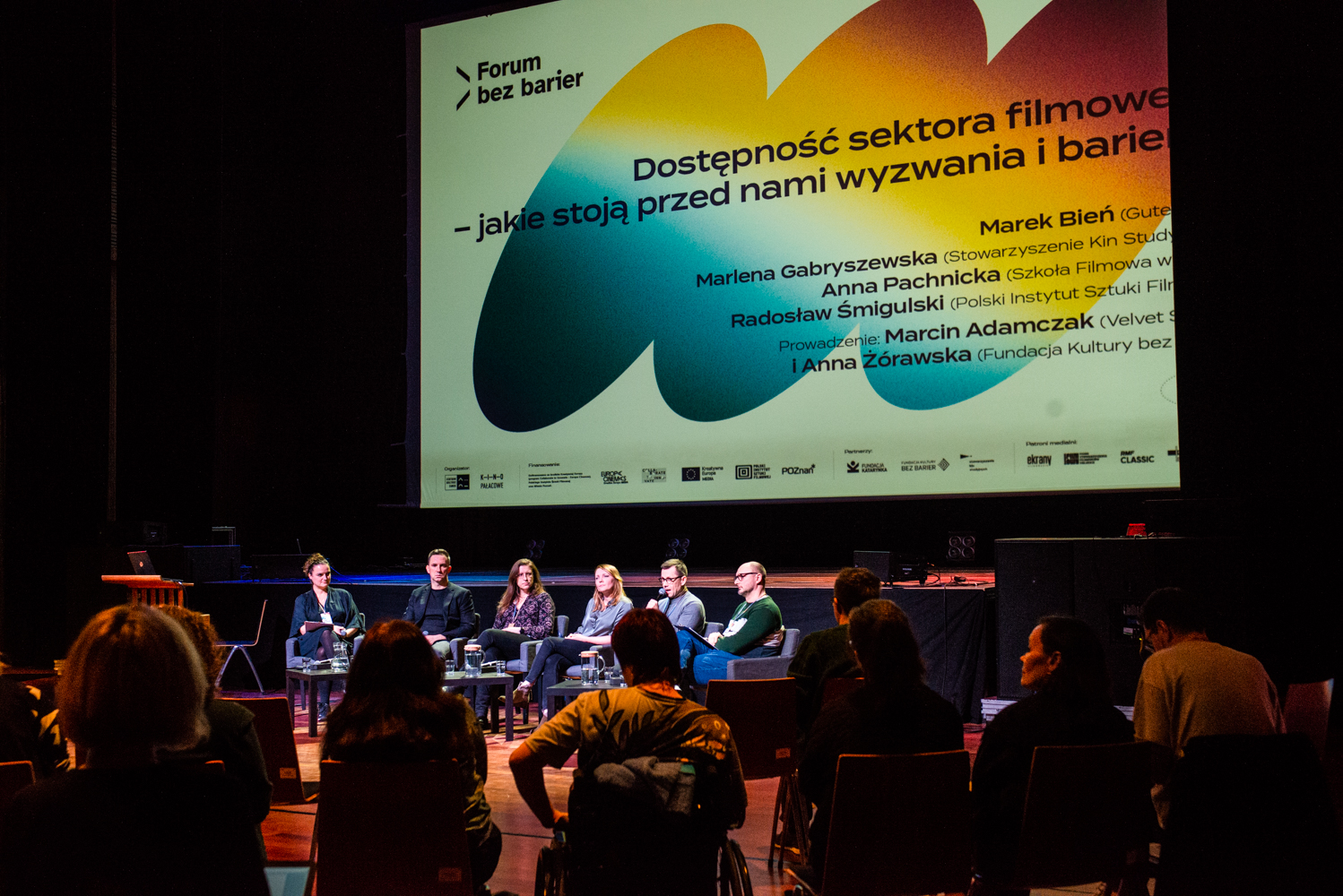 Sześć osób siedzi na scenie w Sali Wielkiej CK Zamek i uczestniczą w dyskusji. Za nimi slajd z prezentacji pt."Dostępność sektora filmowego - jakie stoją przed nami wyzwania i bariery?".