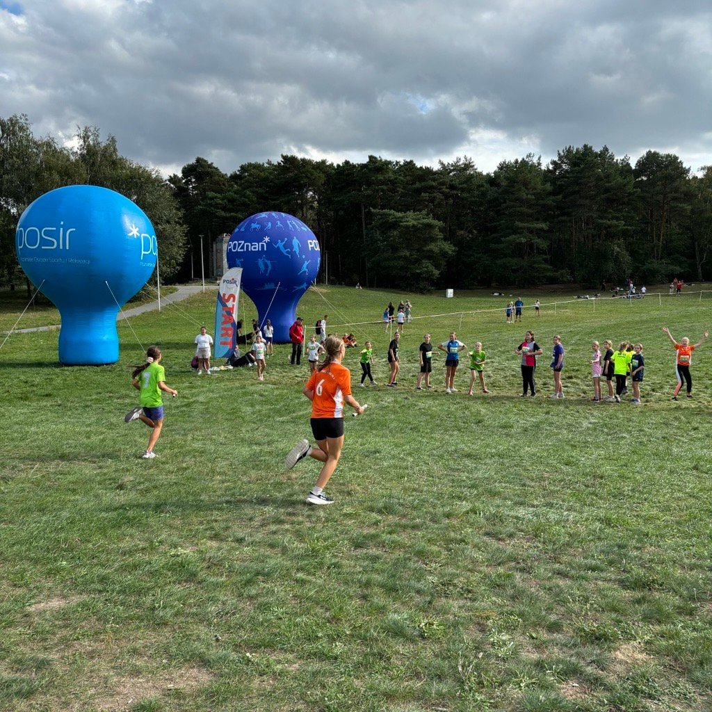 Sztafetowe Biegi Przełajowe - na polanie biegną dziewczynki, które szykują się do przekazania pałeczek kolejnym zawodniczkom. W tle widać las
