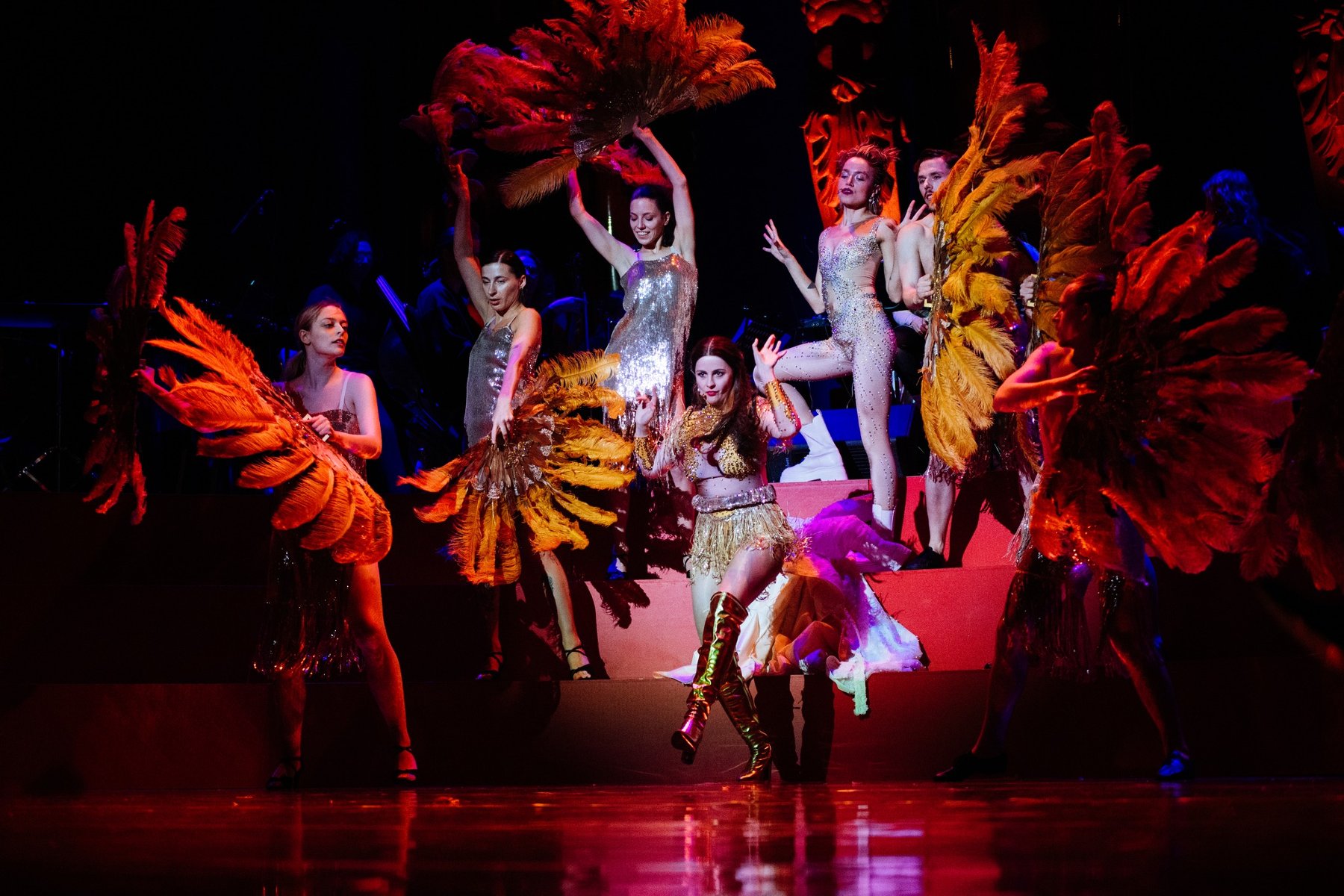 Tancerki w błyszących strojach i wielkimi wahlarzami ze słotych piór. W centrum w złotym kostiumie gwiazda wieczoru - młoda kobieta.