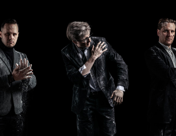 Trzech mężczyzn w ciemnych garniturach i marynarkach pozuje na czarnym tle. Ich ubrania, twarze i dłonie są mocno pobrudzone białym proszkiem przypominającym mąkę lub pył. Mężczyzna po lewej klaszcze w dłonie, wzbijając chmurę pyłu, środkowy otrzepuje ramię, a ten po prawej podwija rękaw.