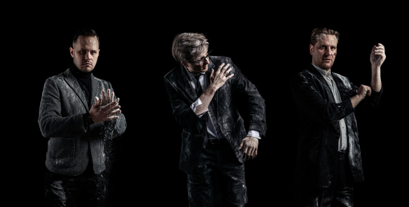 Trzech mężczyzn w ciemnych garniturach i marynarkach pozuje na czarnym tle. Ich ubrania, twarze i dłonie są mocno pobrudzone białym proszkiem przypominającym mąkę lub pył. Mężczyzna po lewej klaszcze w dłonie, wzbijając chmurę pyłu, środkowy otrzepuje ramię, a ten po prawej podwija rękaw.