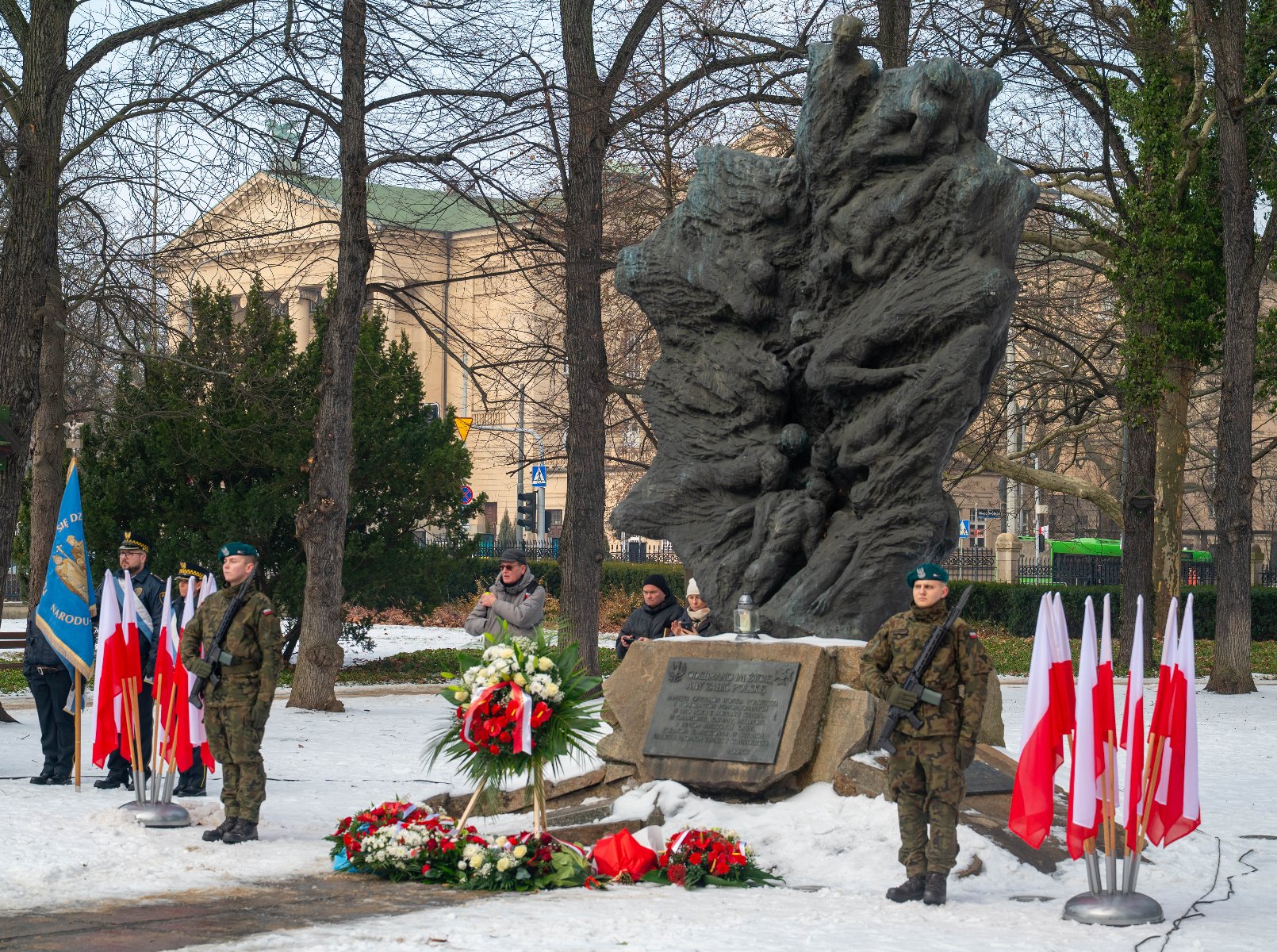 Uroczystość patriotyczna przed pomnikiem w Poznaniu w zimowej scenerii. Na pierwszym planie, po obu stronach monumentu, stoją żołnierze w pełnym umundurowaniu z bronią, pełniąc wartę honorową. Przed pomnikiem złożono liczne wieńce z białych i czerwonych kwiatów. Wokół ustawione są polskie flagi na stojakach.