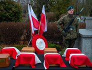 Uroczystość żałobna na cmentarzu. Na pierwszym planie, na stole przykrytym czarnym suknem, ustawiono drewniane urny, z których część przykryta jest biało-czerwonymi materiałami. W tle stoi żołnierz w pełnym umundurowaniu z karabinem, pełniąc wartę honorową. Obok niego znajdują się dwie polskie flagi oraz okrągły wieniec w narodowych barwach.