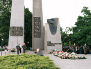 Uroczystości pod Pomnikiem Poznańskiego Czerwca 1956 na placu Mickiewicza. Widoczne są dwa wysokie, betonowe krzyże z datami 1956 oraz 1968, 1970, 1976, 1980 i 1981. Przed pomnikiem płonie znicz, a na ziemi ułożono liczne wiązanki biało-czerwonych kwiatów.