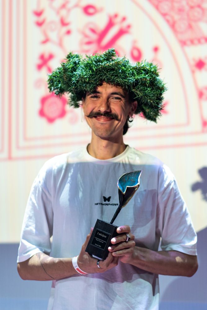 Uśmiechnięty młody mężczyzna trzyma w ręku statuetkę. Na głowie ma zielony wieniec zwycięzcy.