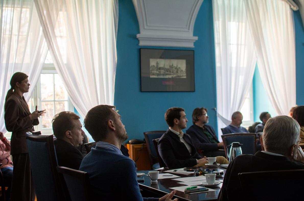 W sali konferencyjnej przy stole siedzi kilka osób, inne osoby siedzą na krzesłach za stołem, jedna osoba stoi i przedstawia prezentację