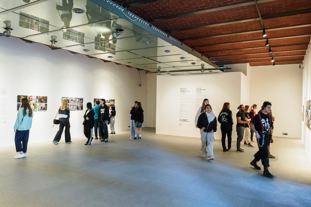 w sali wystawowej około kilknunastu osób ogląda wystawę fotografii, fotografie zawieszone są na białych ścianach