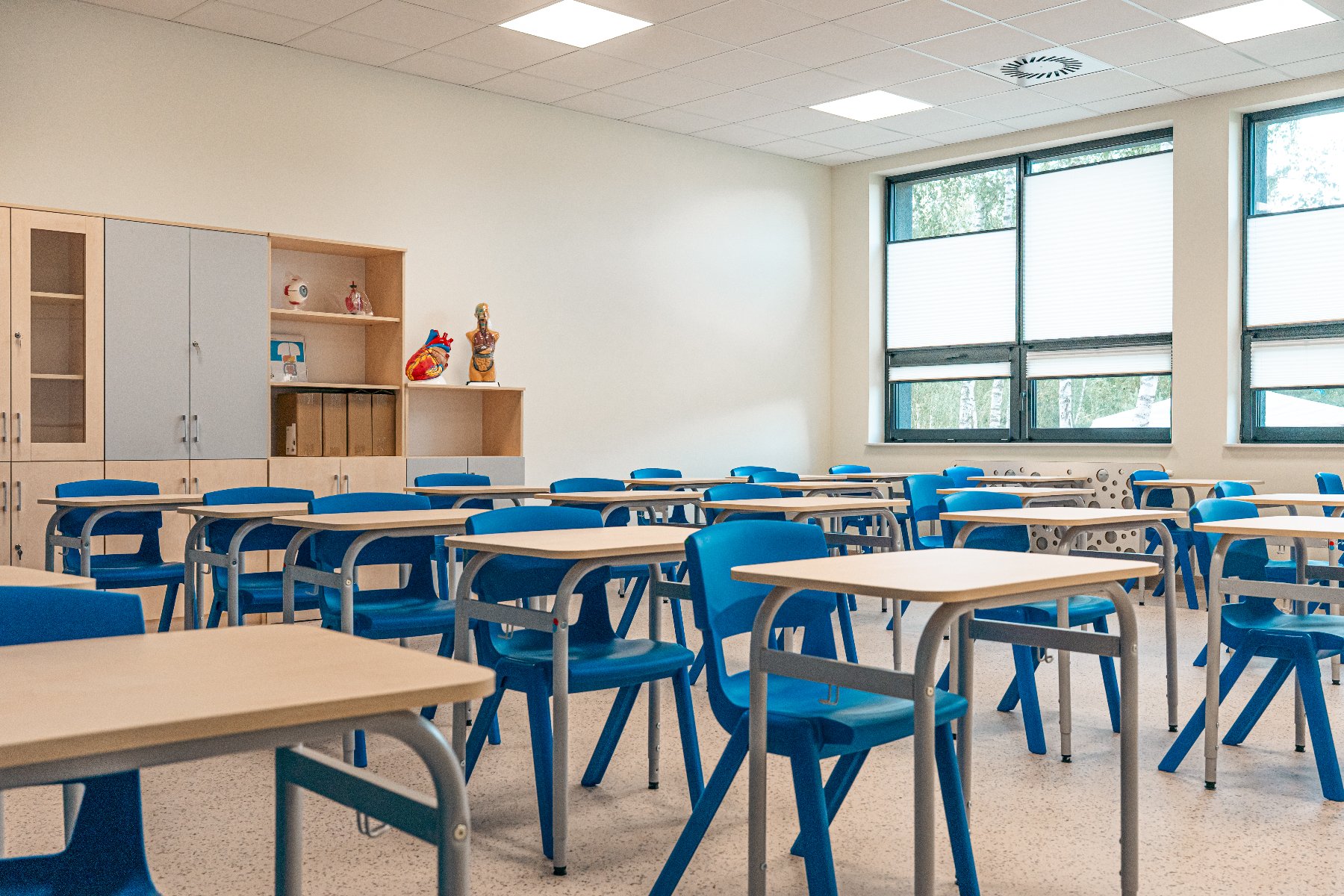 W sali znajdują się rzędy jednoosobowych stolików z jasnymi blatami oraz dopasowane do nich intensywnie niebieskie, plastikowe krzesła na metalowych stelażach.