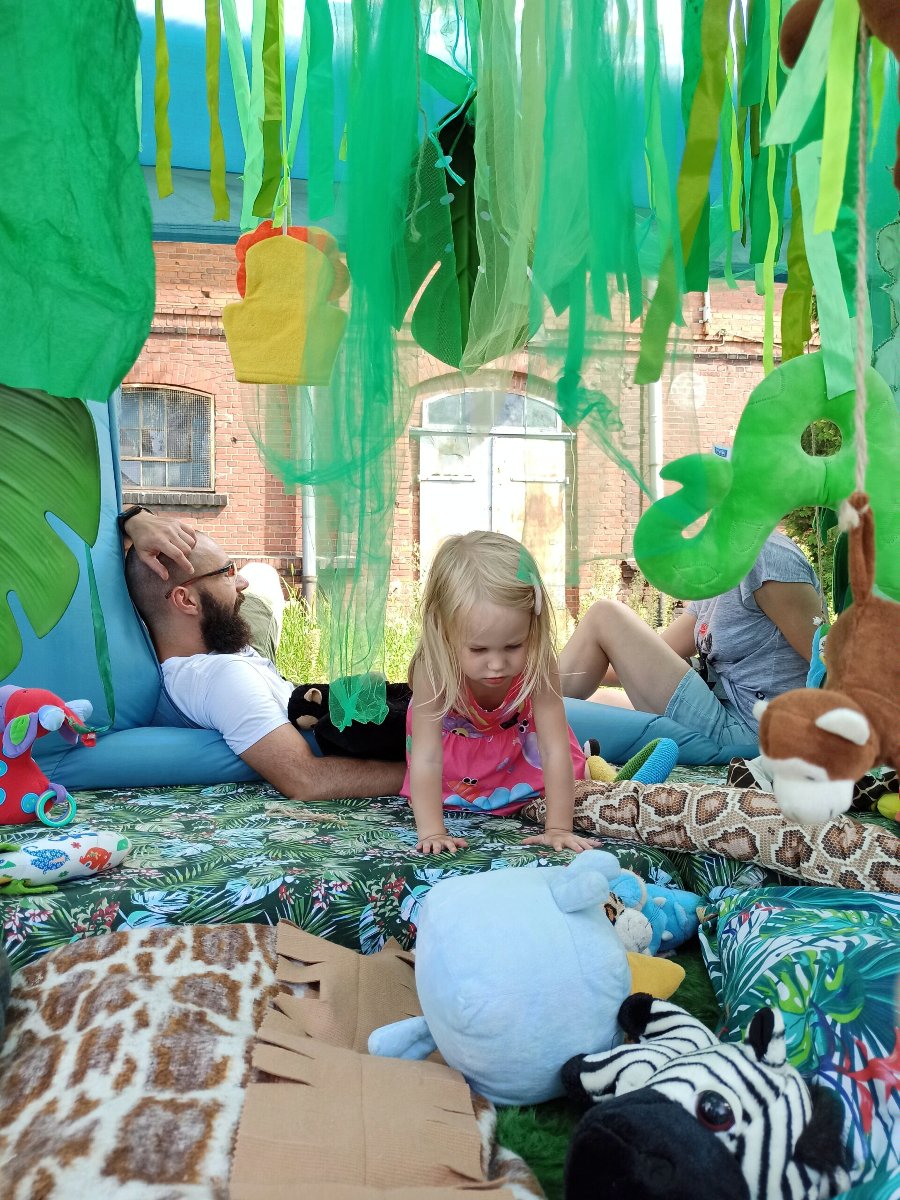 Mała dziewczynka bawi się na kolorowej macie, na której leżą różne pluszaki. Za dziewczyną siedzą rodzice, którzy rozmawiają ze sobą.