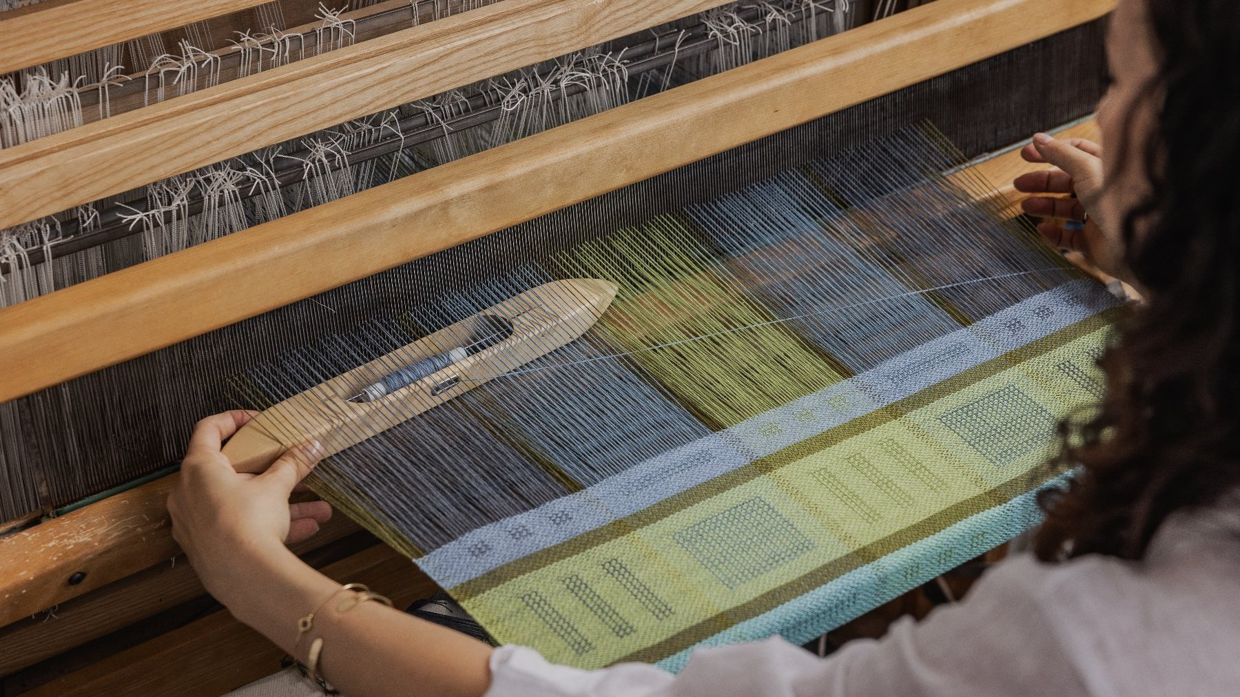 Widoczna od tyłu kobieta o ciemnych, kręconych włosach operuje drewnianym czółenkiem tkackim, przekładając je przez osnowę. Jej lewa dłoń, ozdobiona złotą biżuterią, spoczywa na ramie krosna.