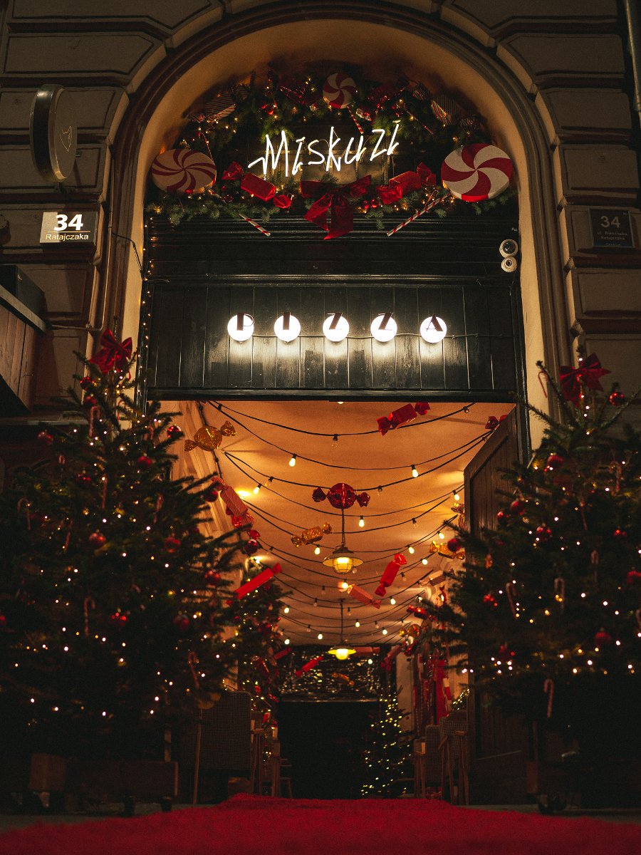 Widok na dwie choinki przystrojone w czerwone ozdoby. Między nimi wejście, nad którym widać rozwieswzone kolorowe lampki z ozdobami.