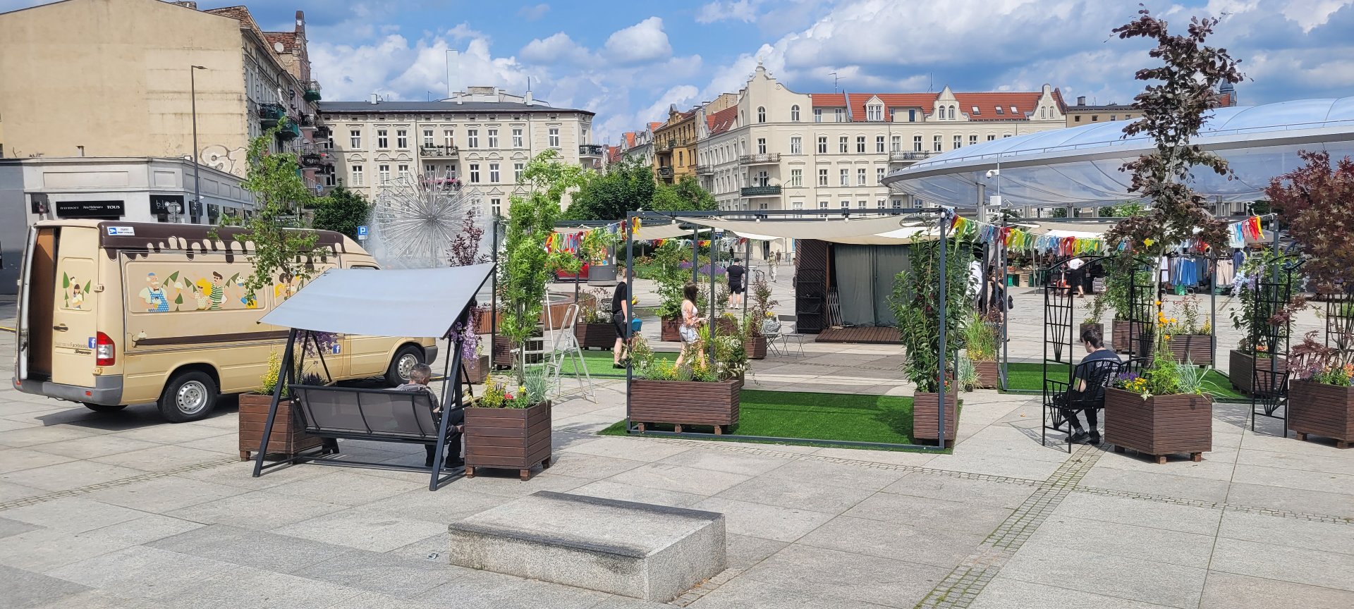 Widok na osoby które rozkładają strefę Łazarskiego Lata. Po lewej stronie bus monterów. Widać rozłożone huśtawki stoliki, baldahim i kolorowe wstążki, które powiewają zawieszone wokół strefy.