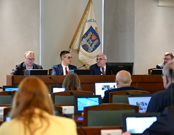 Widok na salę sesyjną podczas posiedzenia. Na podwyższeniu siedzi czterech mężczyzn w garniturach, patrzących przed siebie. W tle wisi biała flaga z herbem i napisem, częściowo widocznym. Na pierwszym planie, rozmazana, widoczna jest publiczność, której część patrzy w ekrany laptopów i monitorów.