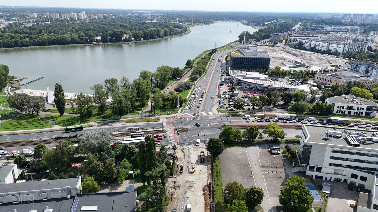 Widok z góry na drogę, która biegnie wzdłuż jeziora. Na zdjęciu widać duży ruch samochodowy na jezdni oraz rondo na skrzyżowaniu. W tle widoczne są budynki mieszkalne, drzewa, a także dźwig.
