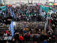 Widok z góry na zatłoczoną halę targową z licznymi stoiskami wędkarskimi. Na pierwszym planie stoisko z rzędami niewielkich akcesoriów w gablotach, przy którym gromadzą się ludzie. W tle widoczne wysokie banery reklamowe, między innymi z napisami "Dystans", "LIBRA LURES" oraz "torqeedo". W głębi hali tłum zwiedzających przemieszczający się między ekspozycjami.