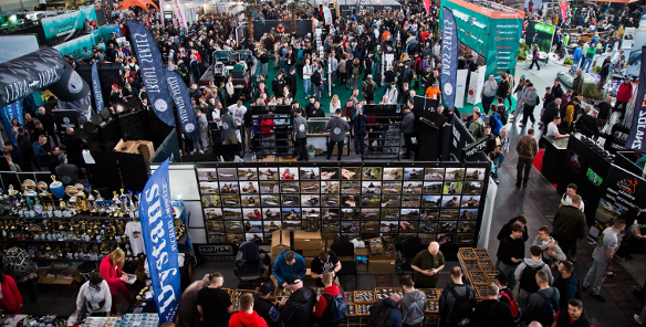 Widok z góry na zatłoczoną halę targową z licznymi stoiskami wędkarskimi. Na pierwszym planie stoisko z rzędami niewielkich akcesoriów w gablotach, przy którym gromadzą się ludzie. W tle widoczne wysokie banery reklamowe, między innymi z napisami "Dystans", "LIBRA LURES" oraz "torqeedo". W głębi hali tłum zwiedzających przemieszczający się między ekspozycjami.
