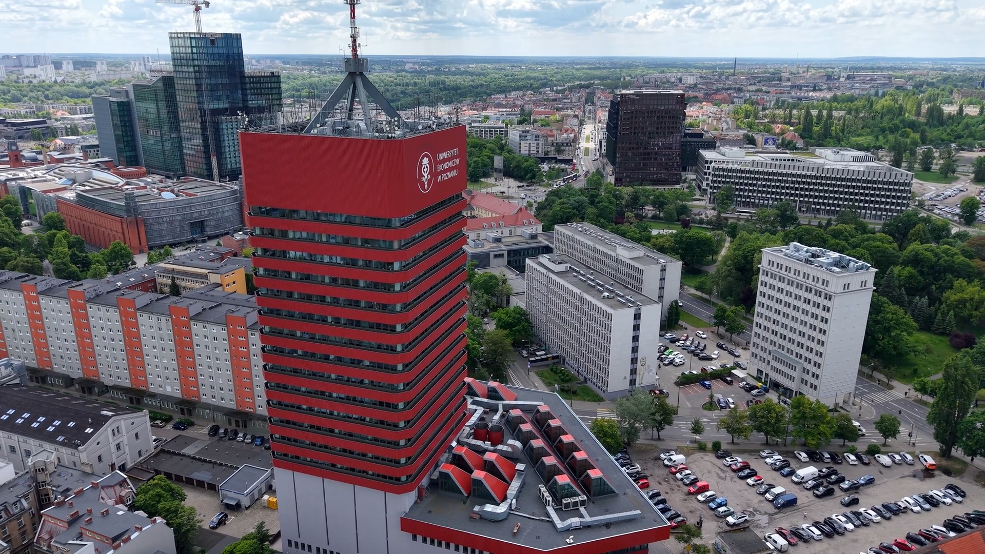 Widok z lotu ptaka na czerwoną wieżę Uniwersytetu Ekonomicznego. Wokół inne budynki w okolicy, zielone drzewa. W tle widok na panoramę Poznania.