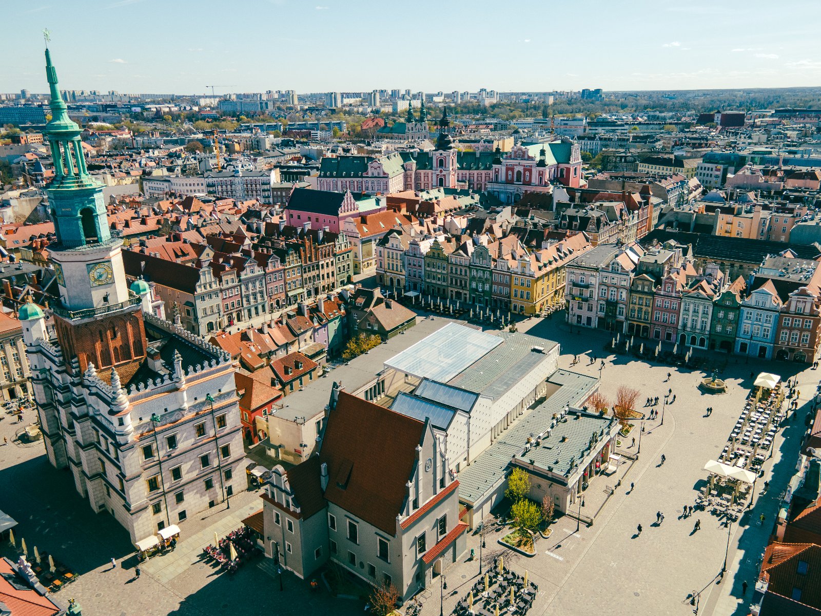 Widok z lotu ptaka na miejski rynek otoczony zabytkowymi kamienicami o kolorowych elewacjach. W centrum placu liczni przechodnie spacerują, a przy stolikach zewnętrznych kawiarni siedzą goście. Charakterystyczną cechą architektury jest wysoka wieża zegarowa z zielonym hełmem.