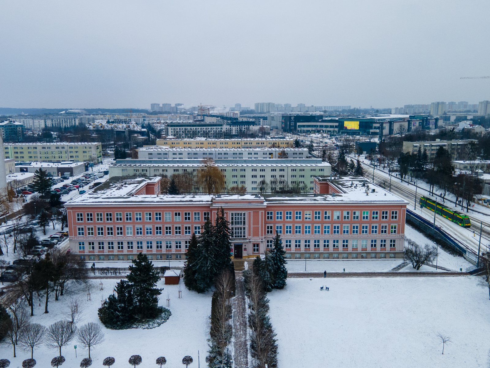 Widok z lotu ptaka na ośnieżone osiedle mieszkaniowe w Poznaniu. W centralnej części znajduje się duży, różowy budynek szkolny w kształcie litery U, przed którym rosną wysokie iglaki. Po prawej stronie widać zielono-żółty tramwaj jadący wzdłuż torowiska, a w tle nowoczesne centrum handlowe z dużym, żółtym ekranem reklamowym pod szarym, zimowym niebem.