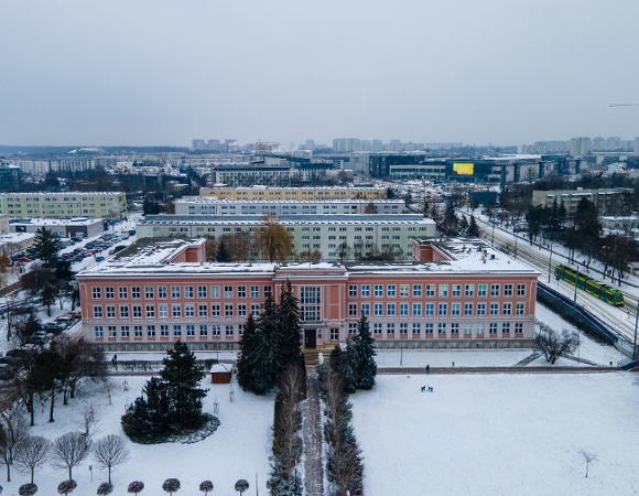 Widok z lotu ptaka na ośnieżone osiedle mieszkaniowe w Poznaniu. W centralnej części znajduje się duży, różowy budynek szkolny w kształcie litery U, przed którym rosną wysokie iglaki. Po prawej stronie widać zielono-żółty tramwaj jadący wzdłuż torowiska, a w tle nowoczesne centrum handlowe z dużym, żółtym ekranem reklamowym pod szarym, zimowym niebem.
