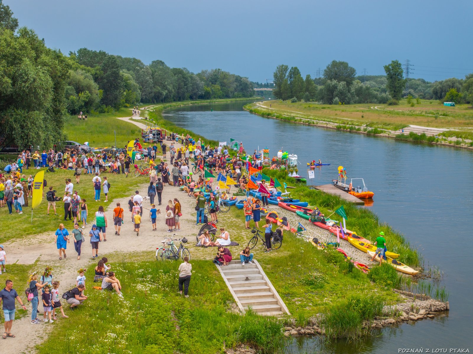 Widok z lotu ptaka na wybrzeże Warty. Na wybrzeżu stoi tłum ludzi w różnym wieku. Przy brzegu zacumowane łódki i kajaki. W tle wije się wstęga Warty otoczona drzewami i trawami.