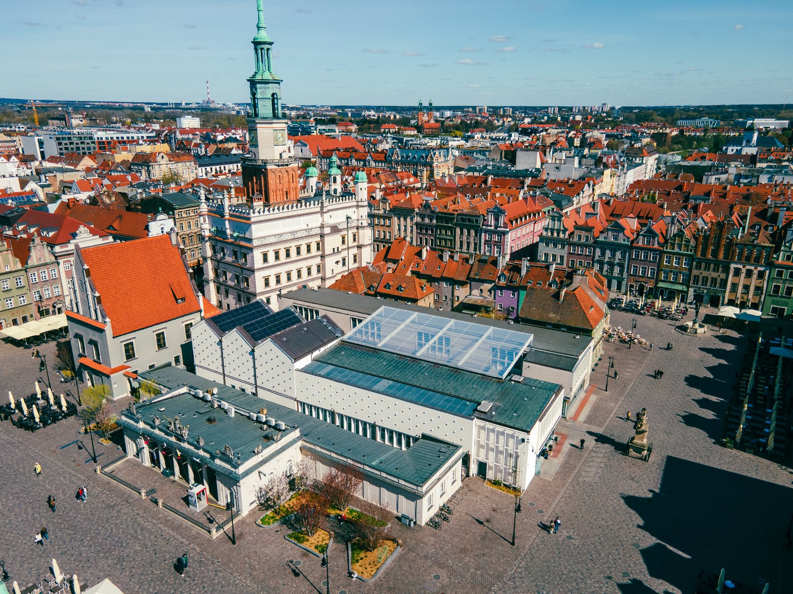 Widok z lotu ptaka na zabytkowe centrum miasta z różnorodną architekturą. W centrum obrazu znajduje się duży budynek z przeszklonym dachem, otoczony kamienicami z czerwonymi dachami. Wyróżnia się także wieża z zielonym hełmem, będąca częścią historycznego obiektu.