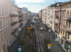 Widok z tóry na ulicy na której trwaja prace budowlane. Teren ulicy jest rozkopany. Widać maszyny budowlane. Po obokach ulicy widac kamienice.