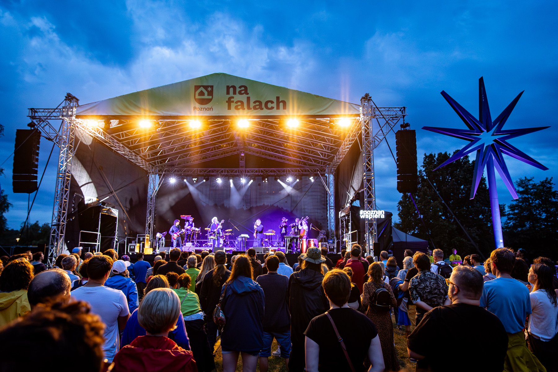 Widownia bawi się na koncercie. Na scenie występuje grupa muzyków, na samej górze widać napis "na falach".