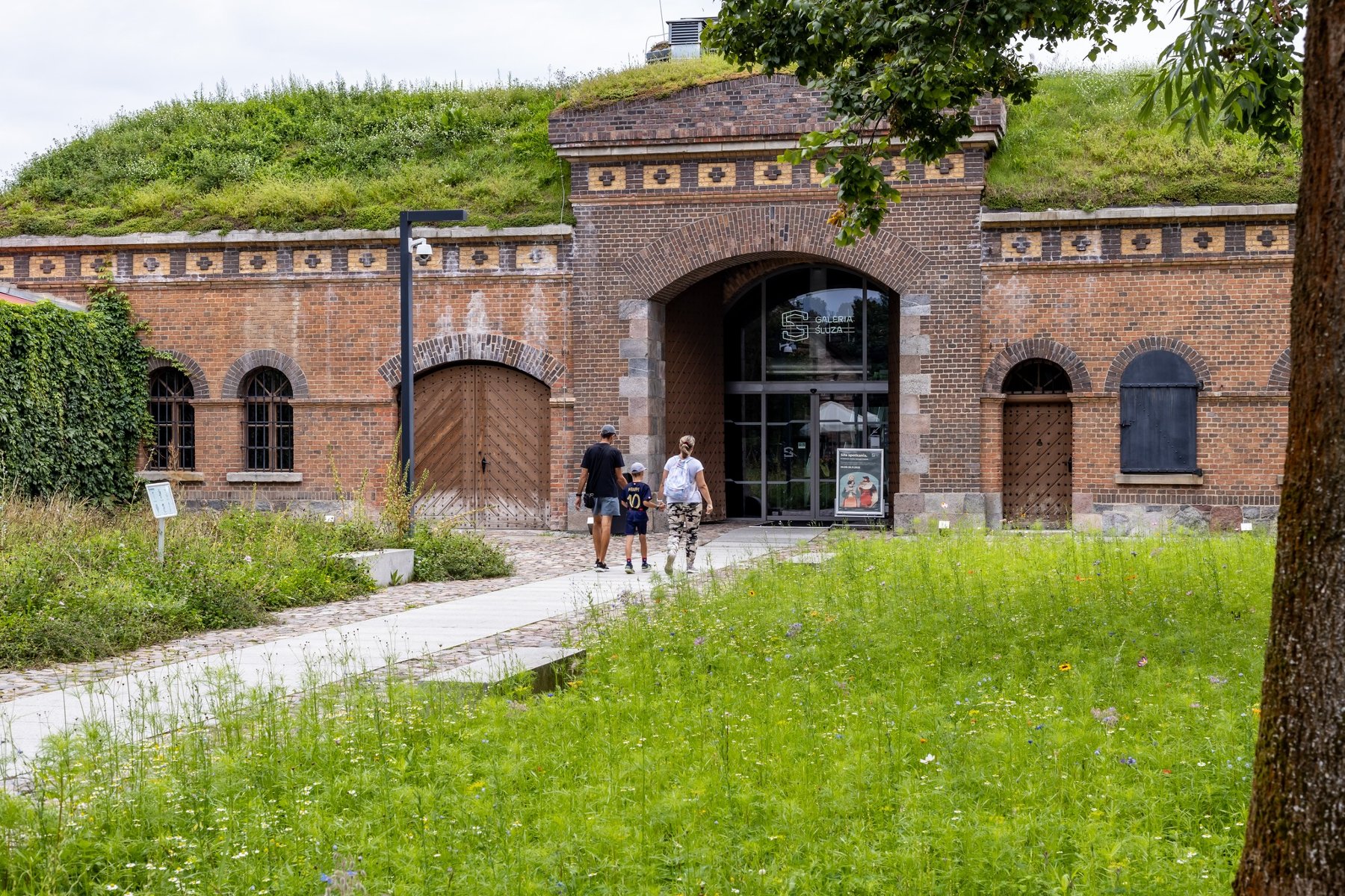 Wieczorny pokaz filmu na świeżym powietrzu na dziedzińcu. Duży ekran wyświetla kolorowy obraz. Przed ekranem, na placu, siedzi liczny tłum ludzi na leżakach i krzesłach, oglądając film. Wokół dziedzińca budynki o czerwonych ścianach i białych zdobieniach.