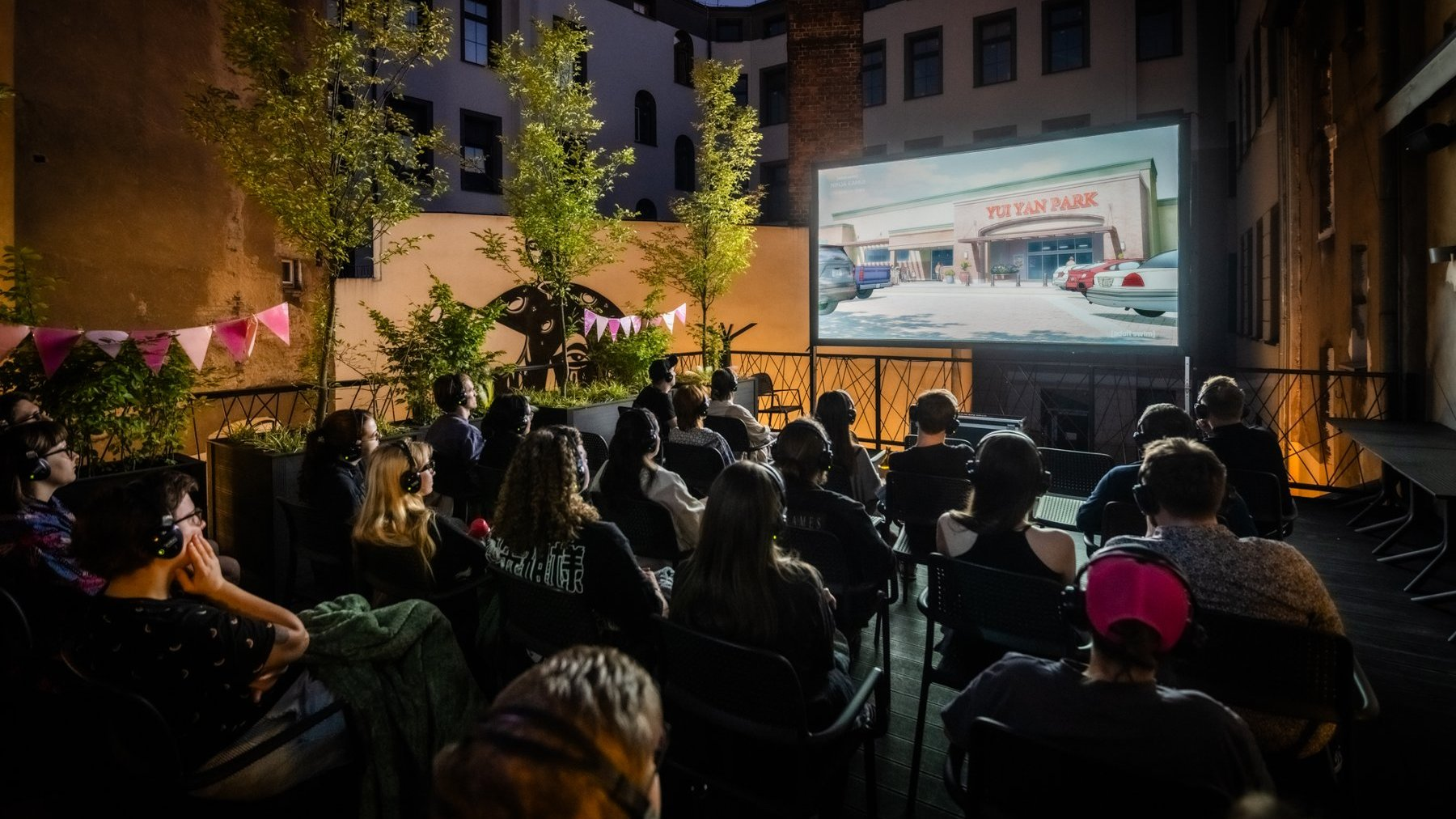 Wieczorny pokaz kina plenerowego. Widzowie siedzą na krzesłach, oglądając film wyświetlany na dużym ekranie. Niektórzy mają na sobie słuchawki. Wokół widoczne są drzewa, lampki i dekoracje w postaci chorągiewek.