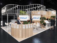 Wizualizacja nowoczesnego stoiska targowego miasta Poznań w ciemnej hali. Konstrukcja stoiska wykonana jest z jasnego drewna i białych, pionowych szczebli tworzących falistą linię u góry. Na stoisku widoczne są białe napisy "POZnan*", ekrany z tym samym logotypem oraz lady ekspozycyjne. Wewnątrz znajdują się stoliki z krzesłami, dekoracje z czerwonych kwiatów oraz zieleń w donicach. Przy ladach i ekranach stoją postacie ludzi w strojach biznesowych.