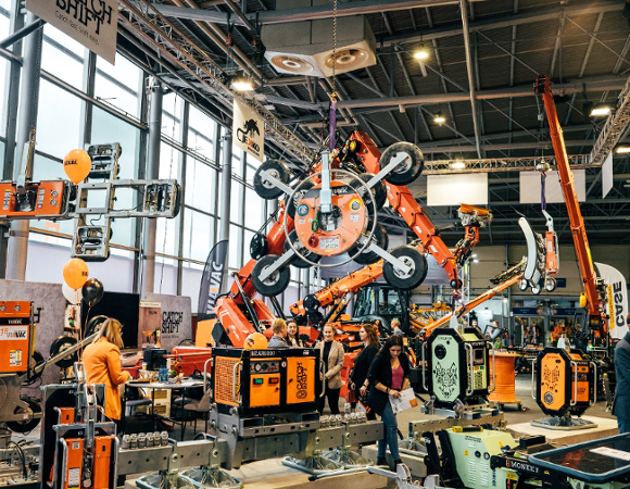 Wnętrze dużej hali targowej z ekspozycją ciężkich maszyn budowlanych i podnośników próżniowych marek VIAVAC oraz CATCH SHIFT. Na pierwszym planie pomarańczowe i zielone urządzenia z grafiką wilka, w głębi ramię dźwigu trzymające duży chwytak z przyssawkami. Wśród maszyn spacerują odwiedzający, widoczne są również pomarańczowe i czarne balony.