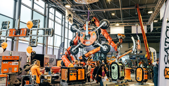 Wnętrze dużej hali targowej z ekspozycją ciężkich maszyn budowlanych i podnośników próżniowych marek VIAVAC oraz CATCH SHIFT. Na pierwszym planie pomarańczowe i zielone urządzenia z grafiką wilka, w głębi ramię dźwigu trzymające duży chwytak z przyssawkami. Wśród maszyn spacerują odwiedzający, widoczne są również pomarańczowe i czarne balony.