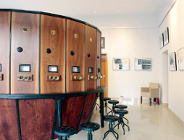 Wnętrze jasnego pomieszczenia z dużym, okrągłym fotoplastikonem wykonanym z ciemnego drewna. Urządzenie posiada ponumerowane stanowiska z mosiężnymi wizjerami, przy których ustawiono czarne, obrotowe taborety. Na białych ścianach w tle wiszą oprawione fotografie i plakaty, a w głębi stoi pojedynczy beżowy fotel.