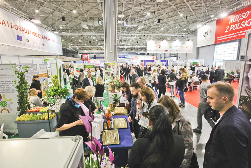 Wnętrze pawilonu targowego. Widać na nim stoiska handlowe i tłumy ludzi.