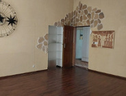 Wnętrze pokoju w mieszkaniu z nietypowymi dekoracjami. Ściany są pomalowane na kolor żółty, a wokół futryny drzwi ułożono nieregularną kompozycję z ozdobnego, piaskowego kamienia. Po lewej stronie wisi duża, ażurowa dekoracja ścienna w kształcie słońca lub zegara, a po prawej obraz nawiązujący do sztuki starożytnego Egiptu.