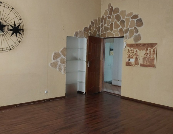 Wnętrze pokoju w mieszkaniu z nietypowymi dekoracjami. Ściany są pomalowane na kolor żółty, a wokół futryny drzwi ułożono nieregularną kompozycję z ozdobnego, piaskowego kamienia. Po lewej stronie wisi duża, ażurowa dekoracja ścienna w kształcie słońca lub zegara, a po prawej obraz nawiązujący do sztuki starożytnego Egiptu.