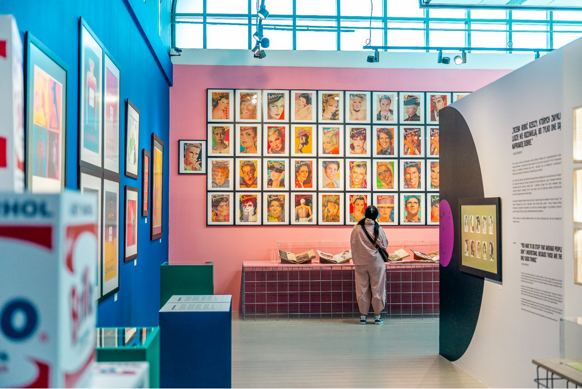 Wnętrze sali ekspozycyjnej, widać kobietę przyglądającą się pracom artysty wywieszonym na ścianie.