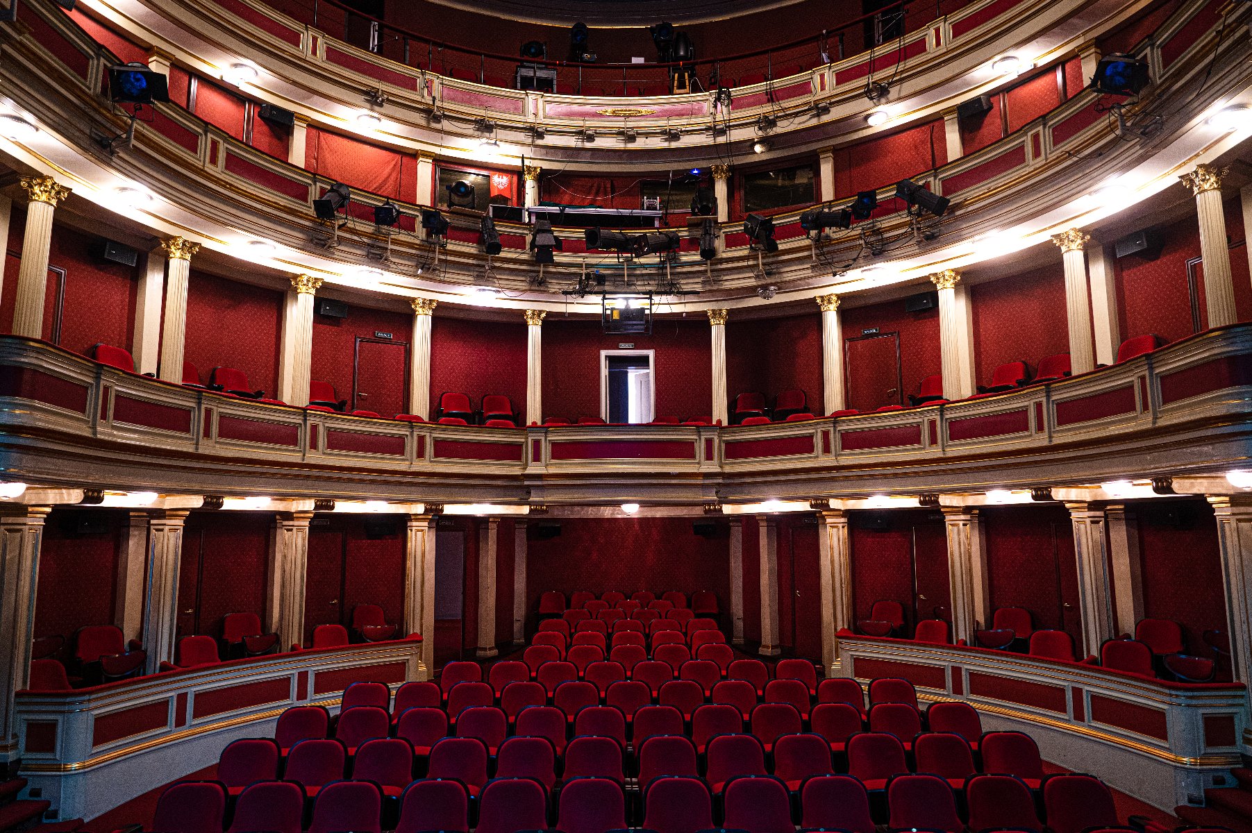 Wnętrze teatru z wieloma poziomami widowni: parter oraz dwa balkony. Fotele obite czerwonym materiałem. Ściany zdobione czerwonymi panelami i złotymi akcentami. Architektura zawiera kolumny i bogate zdobienia, nadając miejscu klasyczny i elegancki wygląd.
