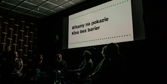 Wnętrze zaciemnionej sali kinowej. Na dużym ekranie widnieje czarny napis na białym tle: »Witamy na pokazie Kina bez barier«, ujęty w dwie poziome linie z zielonych kropek. Poniżej ekranu, w półmroku, siedzi grupa osób odwrócona tyłem lub bokiem do obiektywu, patrząca w stronę wyświetlanego komunikatu.
