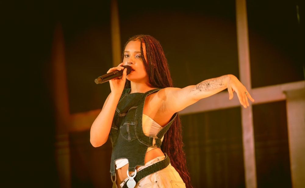 Wokalistka opiera twarz na dłoniach, jej oczy są zamyślone. Ma na sobie białą sukienkę jak mgiełka, a każdy jej paznokieć jest w innym kolorze.