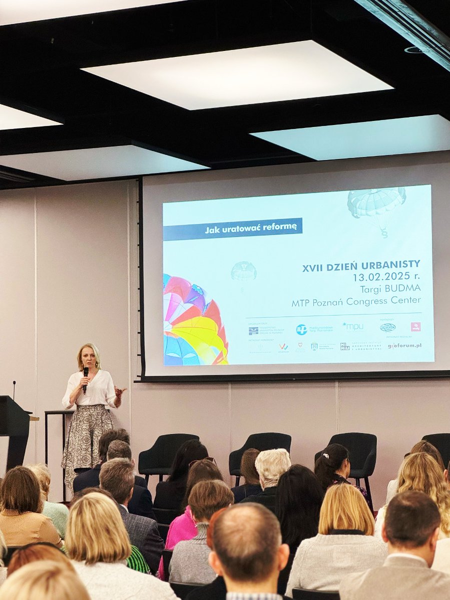 Wystąpienie na konferencji lub seminarium. Osoba przy mównicy mówi do mikrofonu, za nią siedzi trzech uczestników na scenie, słuchających uważnie. Za mównicą znajduje się duży ekran z tekstem i logami.