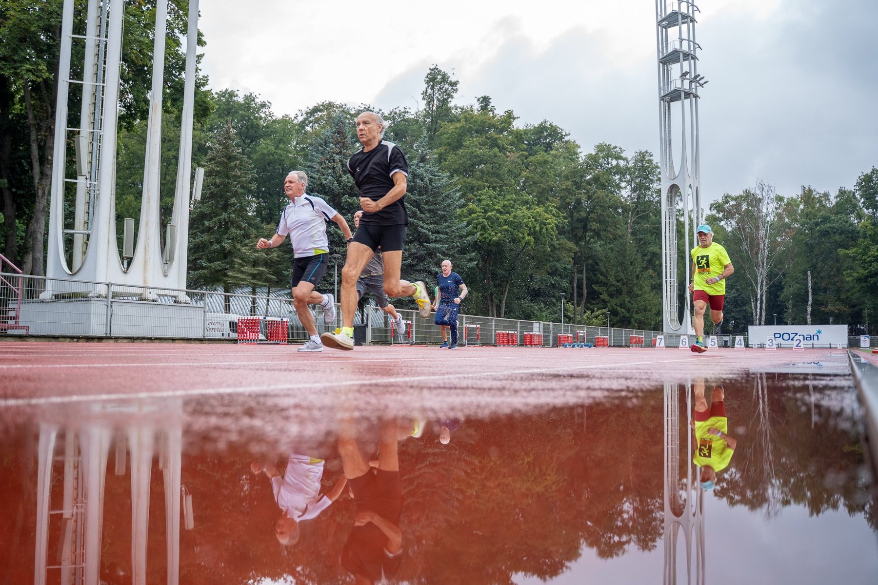 XVIII Spartakiada Seniorów za nami, fot. Agnieszka Wojt-Kaczmarek - 4 seniorów biegnie po mokrej bieżni, w tle widać białe jupitery oraz las