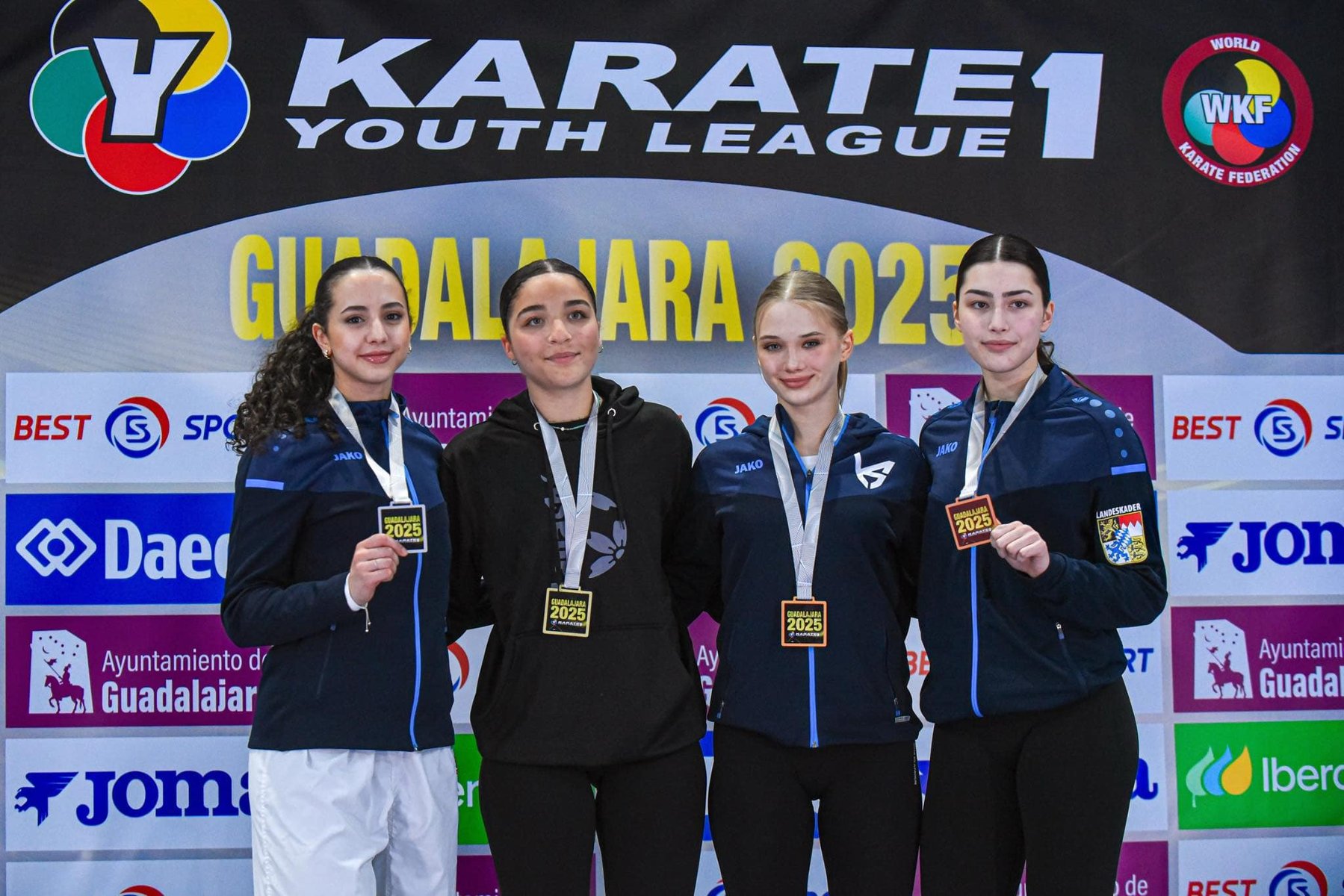 Zawodniczka Kuzi Sport na podium z medalem na zawodach Karate 1 - Youth League, fot. Kuzi Sport - na podium stoją 4 młode kobiety w strojach sportowych. Jedna ma blond włosy, a 3 ciemne. Wszystki mają na szyjach zawieszone medale. W tle widać ściankę zawodów