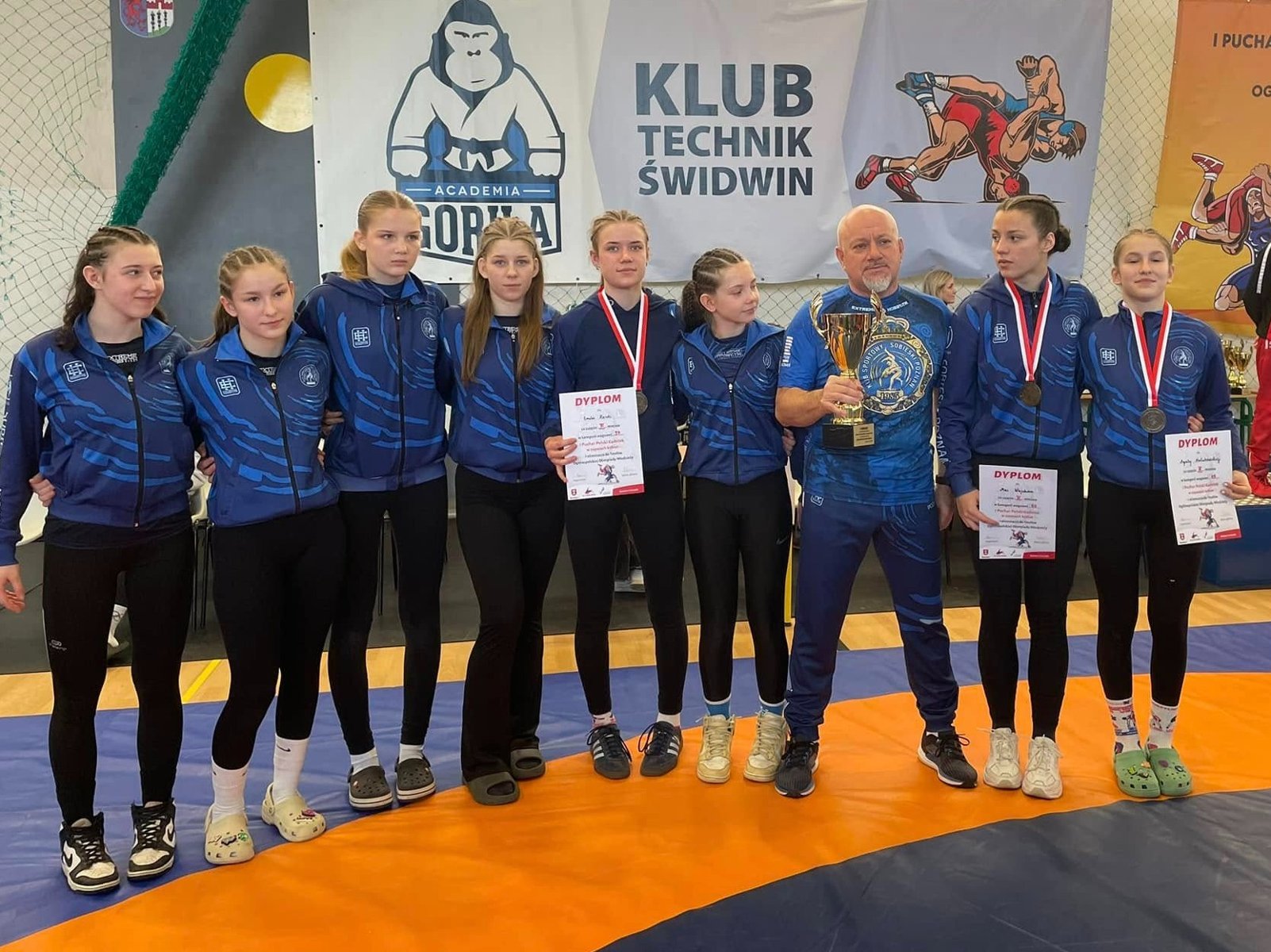 Zawodniczki Sobieskiego Poznań z medalami Pucharu Polski Kadetek w zapasach, fot. Maciej Kula - w strojach klubowych stoją zawodniczki Sobieskiego Poznań, 3 z nich mają medale i dyplomy. W środku stoi trener, który trzyma puchar.
