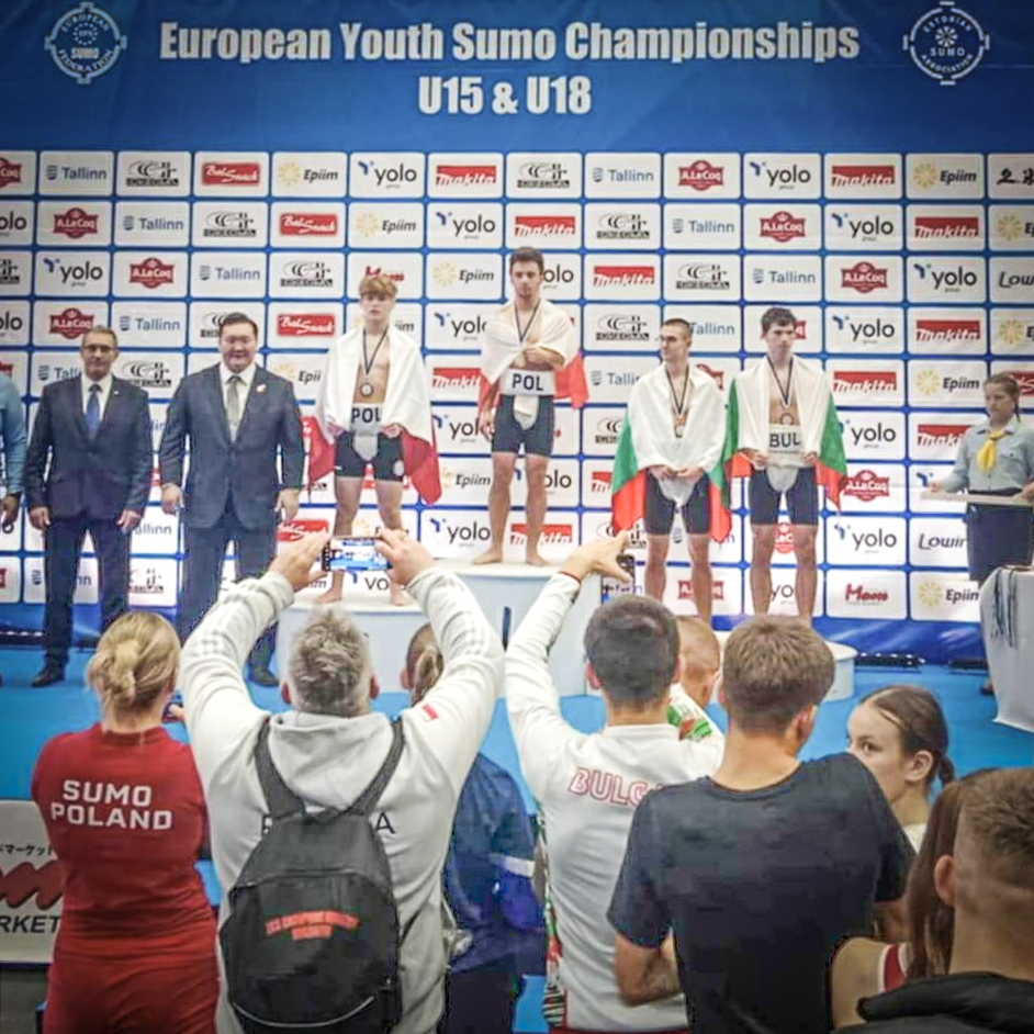 Zawodnik Sobieskiego na najwyższym stopniu podium w Mistrzostwach Europy juniorów w sumo, fot. Maciej Kula - na podium stoi czterech zawodników w półnagich strojach, dwóch ma polskie flagi, a dwóch bułgarskie. Oprócz nich widać wręczających nagrody oficjeli oraz członków ekip robiących zdjęcia