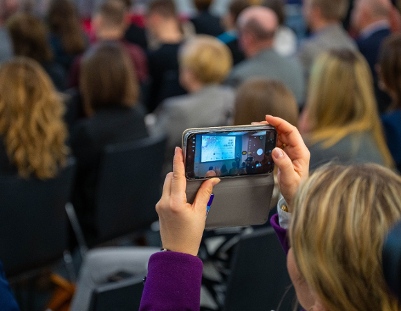 Zbliżenie na dłonie osoby trzymającej smartfon i nagrywającej prezentację podczas konferencji. Na ekranie telefonu widoczny jest slajd wyświetlany na dużym ekranie w głębi sali. W tle rozmyte sylwetki licznie zgromadzonych uczestników siedzących w rzędach.