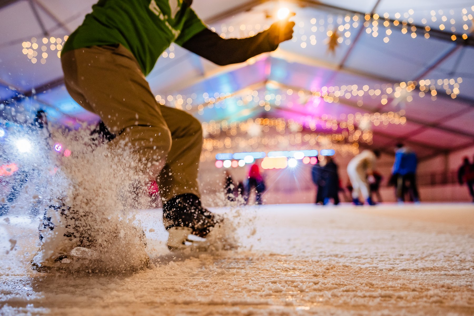 Zbliżenie na nogi i łyżwy osoby w zielonej koszulce, która gwałtownie hamuje na lodowisku, wzbijając chmurę drobnego śniegu. W tle, zamazane, widać innych łyżwiarzy oraz namiot ozdobiony kolorowymi światłami.