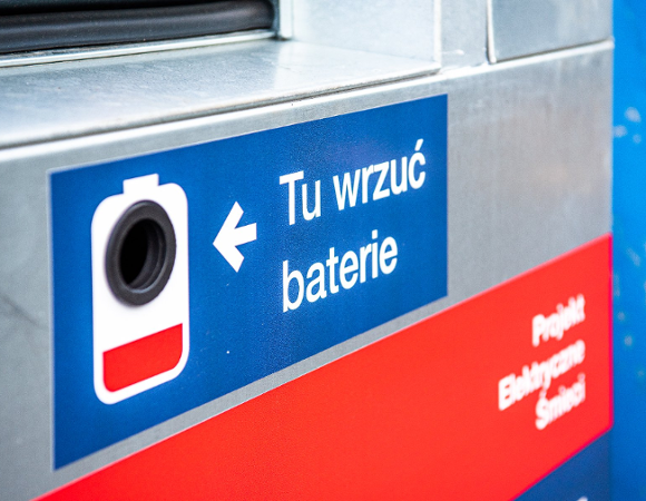 Zbliżenie na otwór wrzutowy pojemnika na odpady elektryczne. Na metalowej obudowie naklejona jest niebieska etykieta z białym napisem »Tu wrzuć baterie«, białą strzałką oraz ikoną baterii. Poniżej znajduje się fragment czerwonej naklejki z napisem »Projekt Elektryczne Śmieci«
